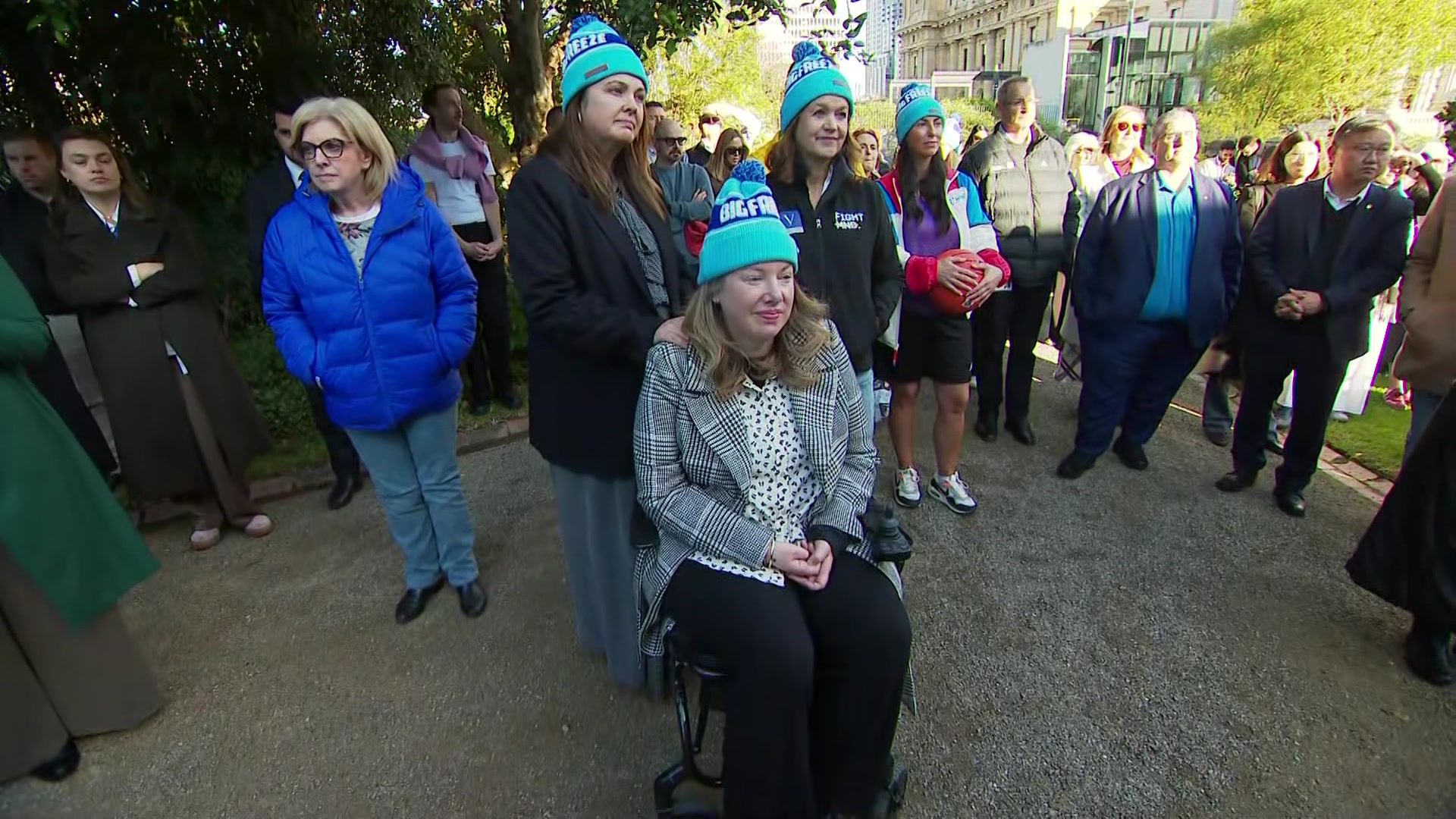 Two women wearing blue beanies stand behind a woman who is seated, wearing the same beanie, near a crowd of people.