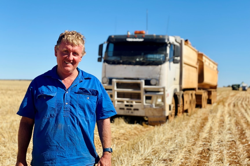 A man wearing a blue shirt stands in a paddock with a grain truck behind him. 