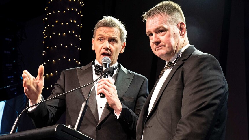 Two middle-aged men stand at a lectern and microphone, with bright lights shining on them, as they wear tuxedos
