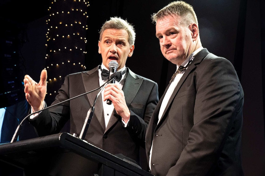 Two middle-aged men stand at a lectern and microphone, with bright lights shining on them, as they wear tuxedos