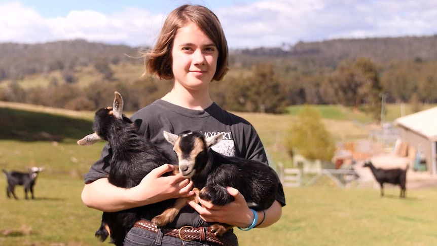 A girl stands outdoors on a farm holding two baby pygmy goats.
