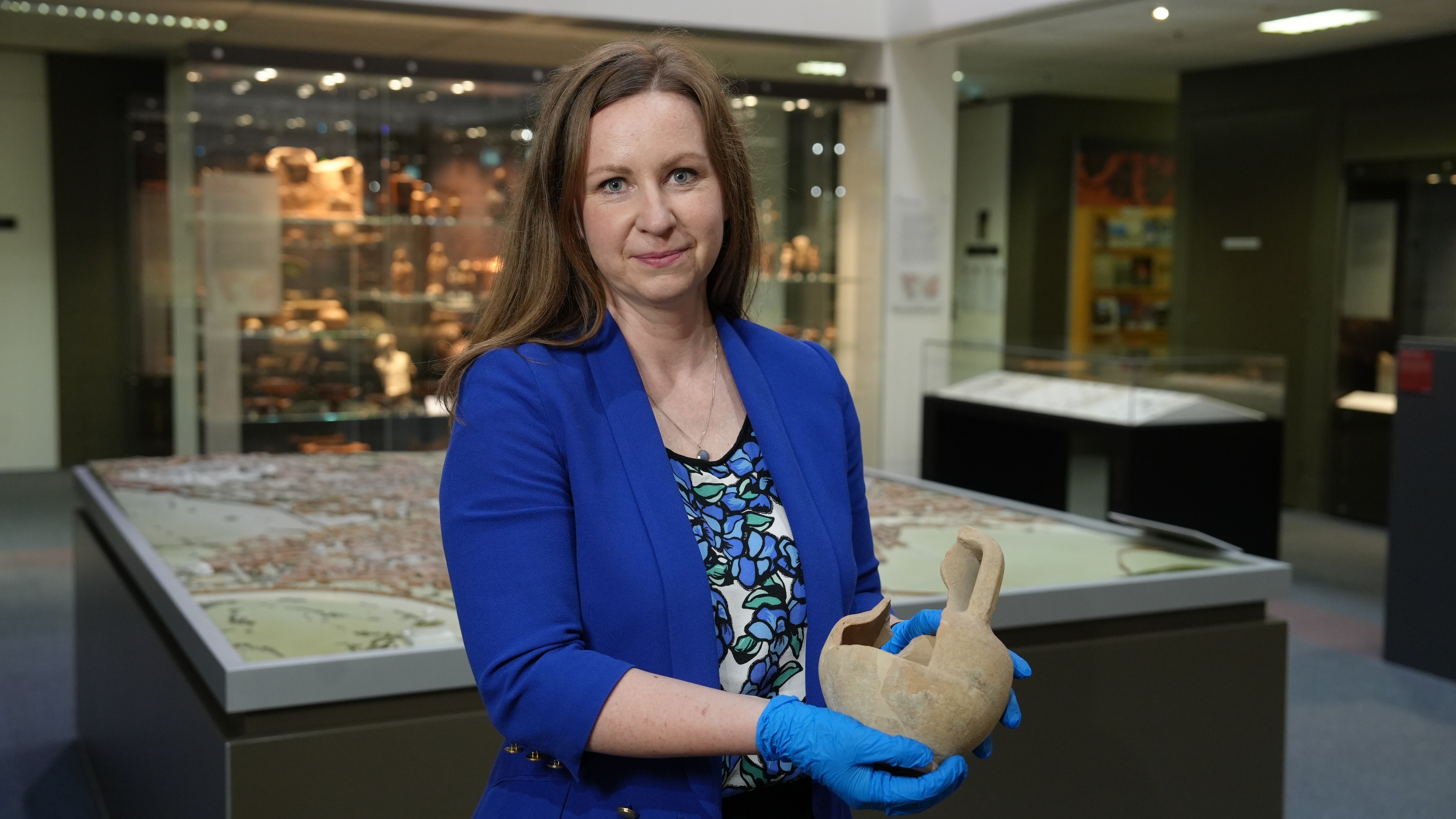 A woman with long brown hair wearing blue latex gloves holds a broken terracotta amphora surrounded by ancient artifacts.