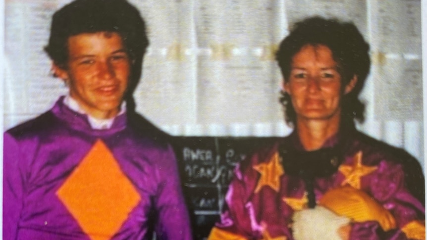 Woman and young man dressed in horse racing silks and holding riding helmet and whip, smiling. 