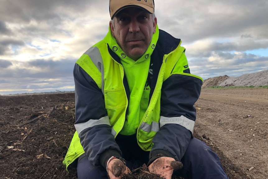 A man in a high-vis jacket kneels on the ground, holding soil in his cupped hands.