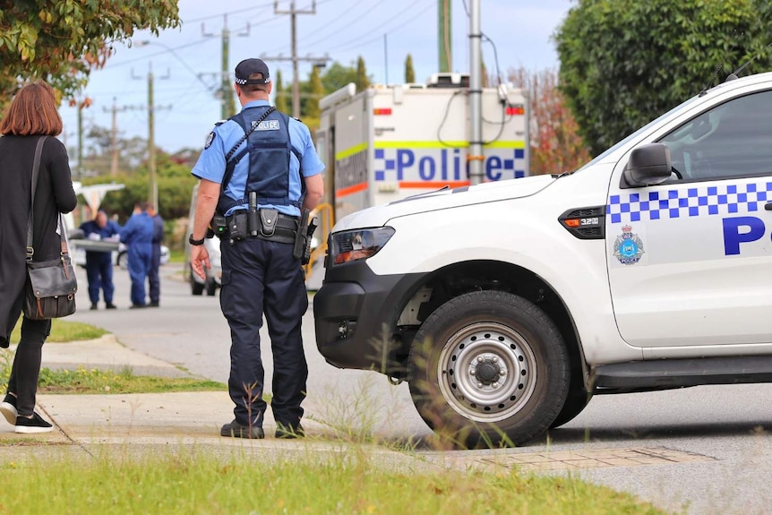 A police officer stands next to a police car blocking off a suburban street with a neighbour looking on.