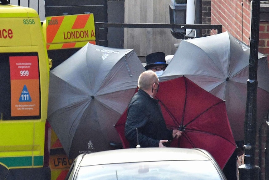 Two men use umbrellas to shield the view between a building and an ambulance door.