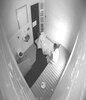 一个穿着长袍的男孩坐在一个有水槽和厕所的牢房里。他的床垫和床单在地板上。