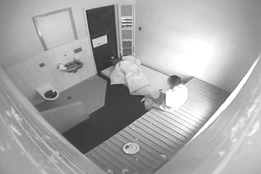 A boy wearing a gown sits in a cell that has a sink and toilet. His mattress and sheet is on the floor.