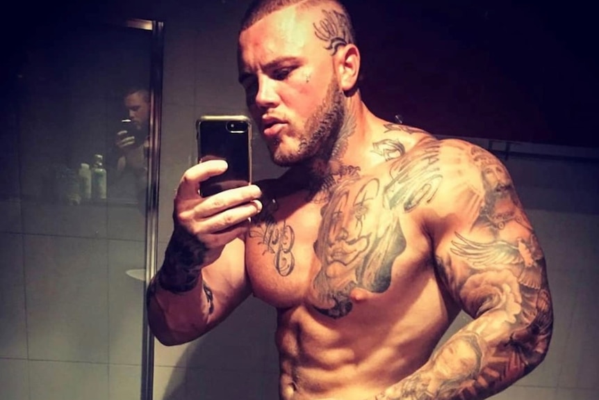Shirtless man with tattoos stands taking a photo in the mirror.