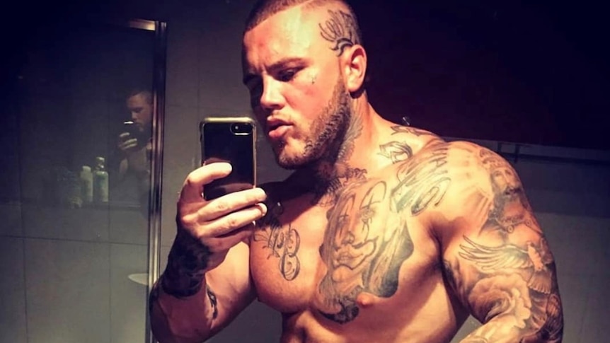 Shirtless man with tattoos stands taking a photo in the mirror.