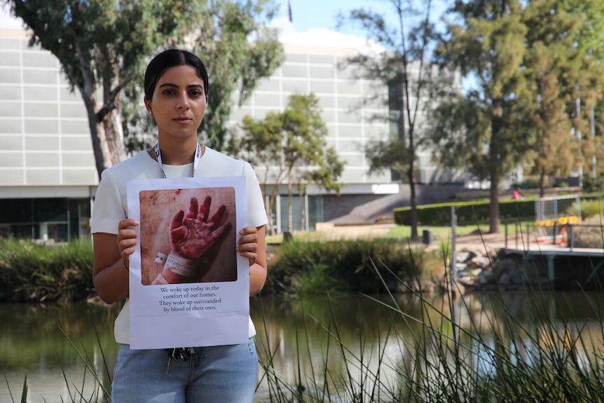 A woman in a white t-shirt stands in front of a pond holding a picture of a bloody hand.