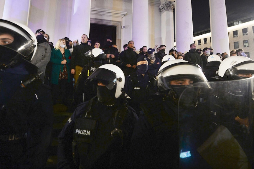 About 100 people stand outside a church with big columns. Police in riot gear stand in front of them.