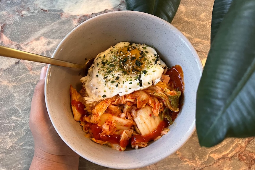 A brown hand is seen holding a bowl sitting on a marble table with a ficus dangling over. The bowl has kimchi, rice and an egg