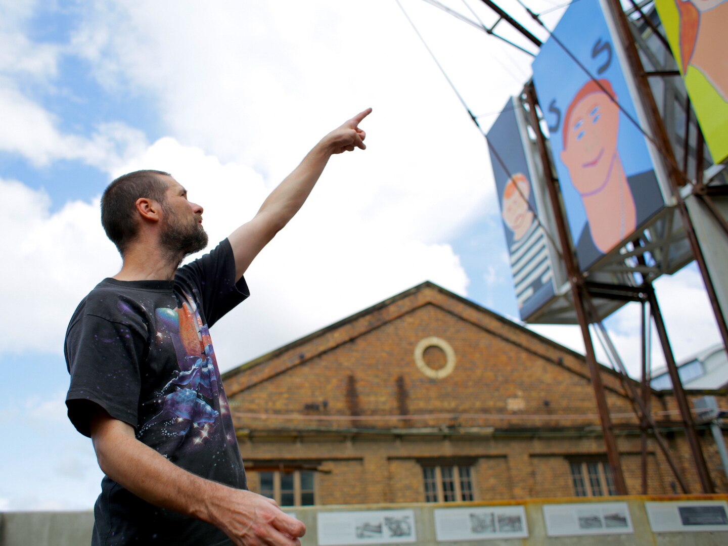 The artist, with short brown hair and a black t-shirt, stands outside pointing at a large billboards with his paintings on it