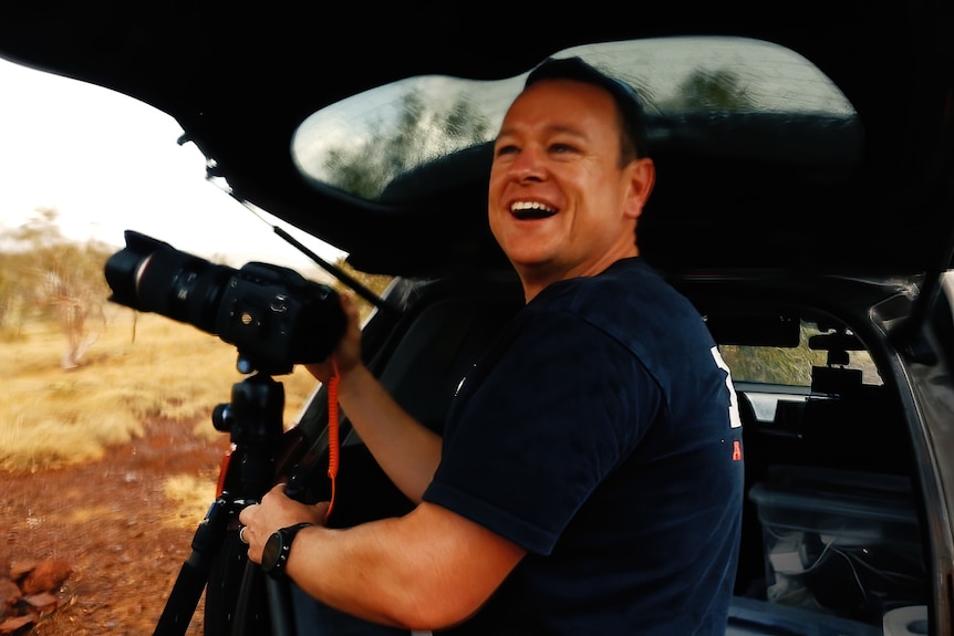 Jordan is excitedly smiling under the cover of his back car door holding his camera.