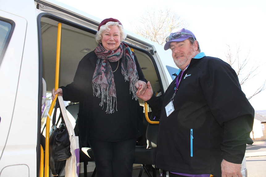 An older woman stepping out of a mini bus helped by a man holding her hand