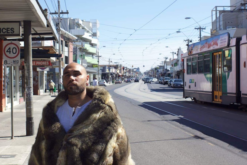 Man stands on road in front of tram with a possum skin cloak around his shoulders