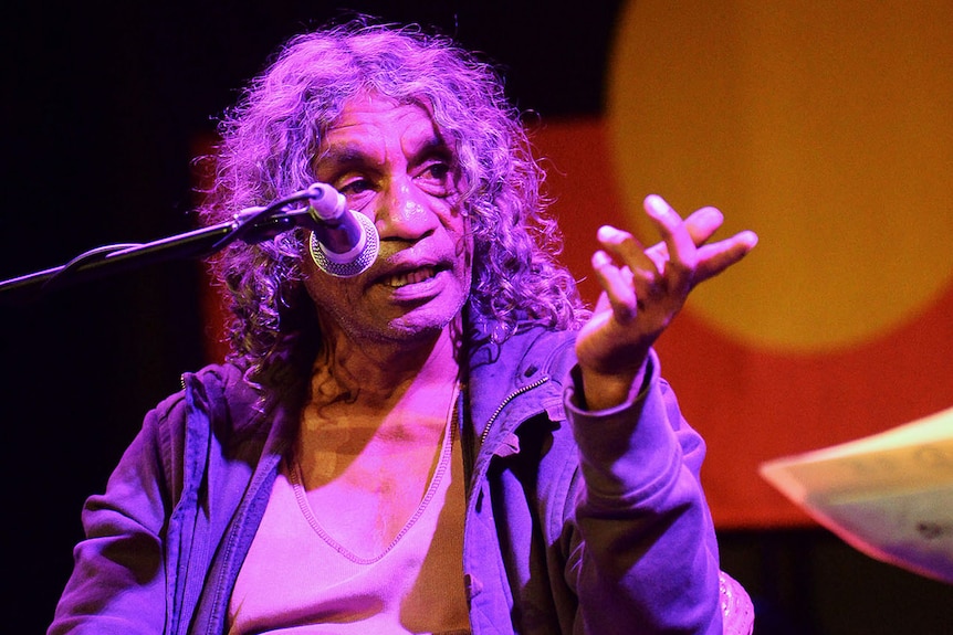 No Fixed Address drummer Bart Willoughby speaks on stage with the Aboriginal flag behind him.