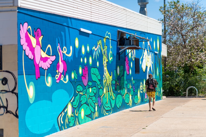 A man walks past the side of a building featuring a colourful mural of dancing animals and plants, on a sunny day.