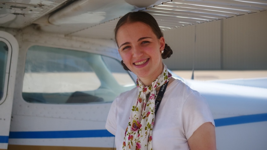 A young woman in silk scarf with her hair in two low buns smiles at the camera as she stands under the wing of a small airplane.