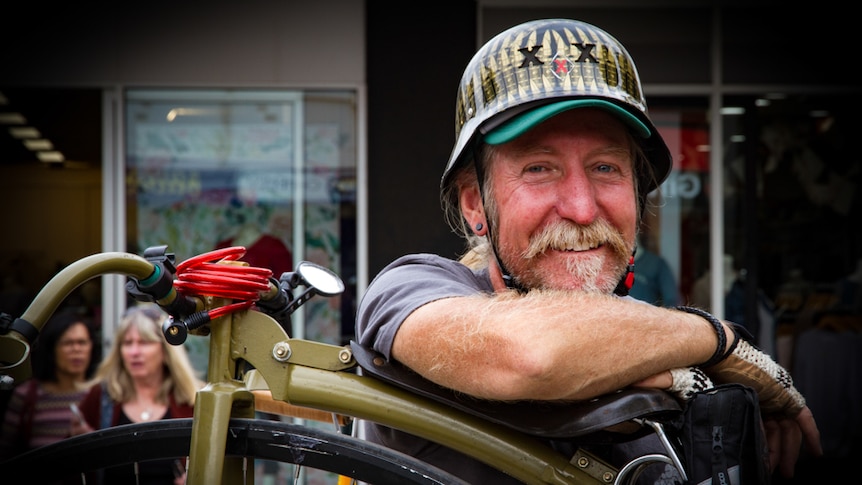 A man wearing a helmet leaning against a penny farthing.