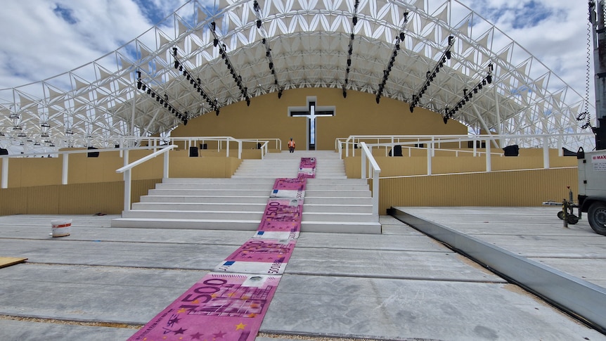 A carpet depicting oversized 500-euro banknotes rolled down the stairs in front of a stage which has a large cross on it.