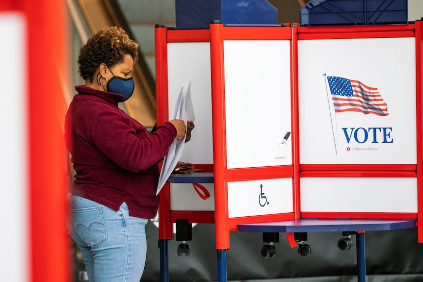 A woman wearing a face mask handles her paper ballot at a booth with the US flag and VOTE emblazoned on it