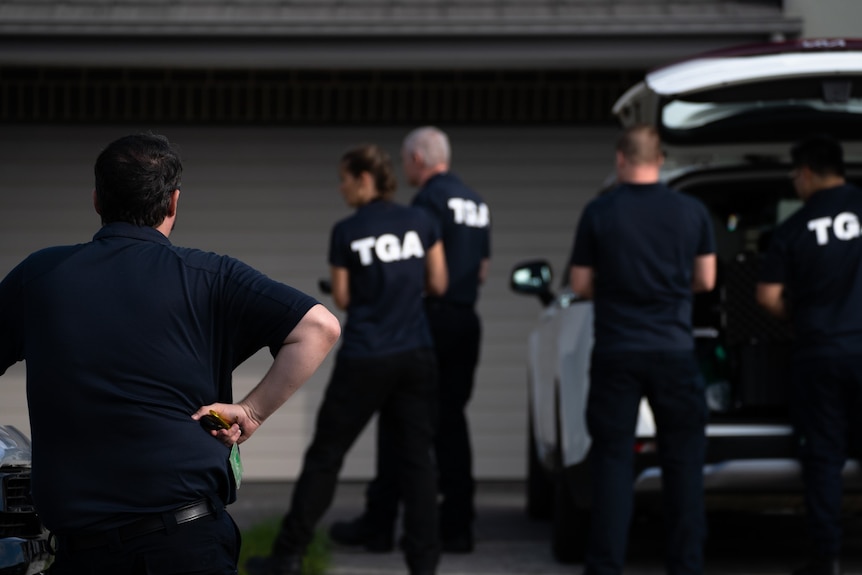 People in polo shirts with TGA on the back stand on the driveway of a house near a car. They are looking away from the camera.