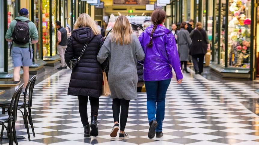 A rear view of three women with linked arms walking in a shopping arcade.