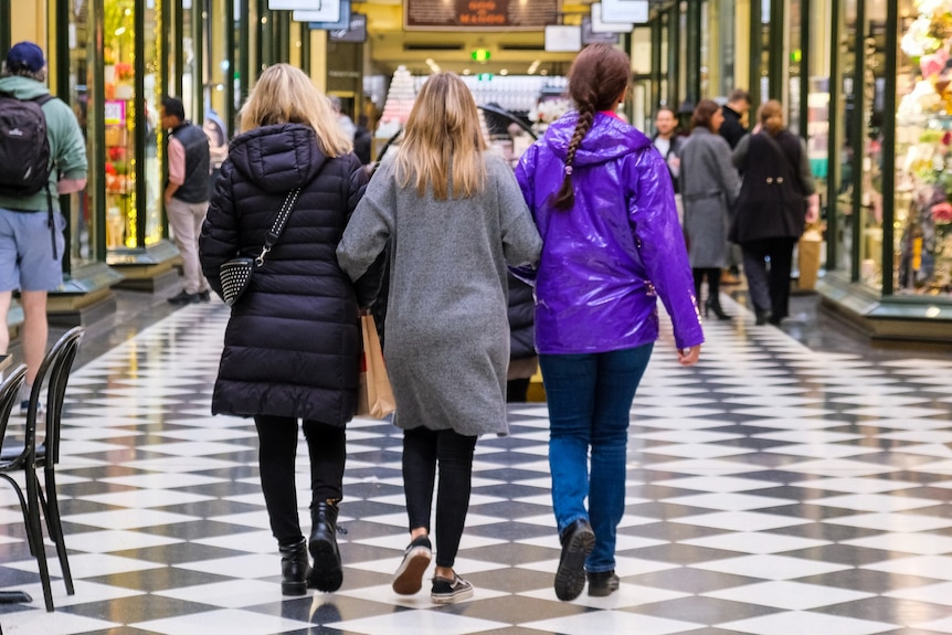 A rear view of three women with linked arms walking in a shopping arcade.