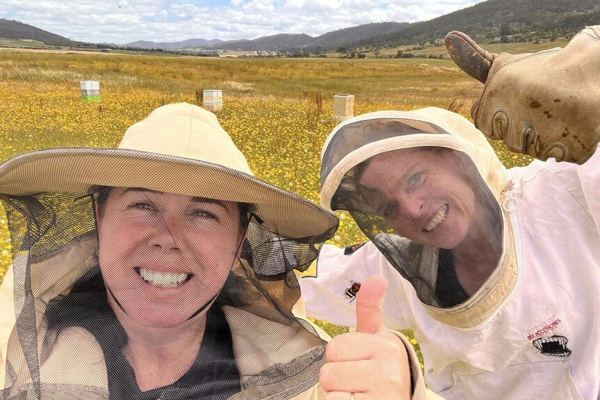 Two women in beekeeping gear smile at the camera.