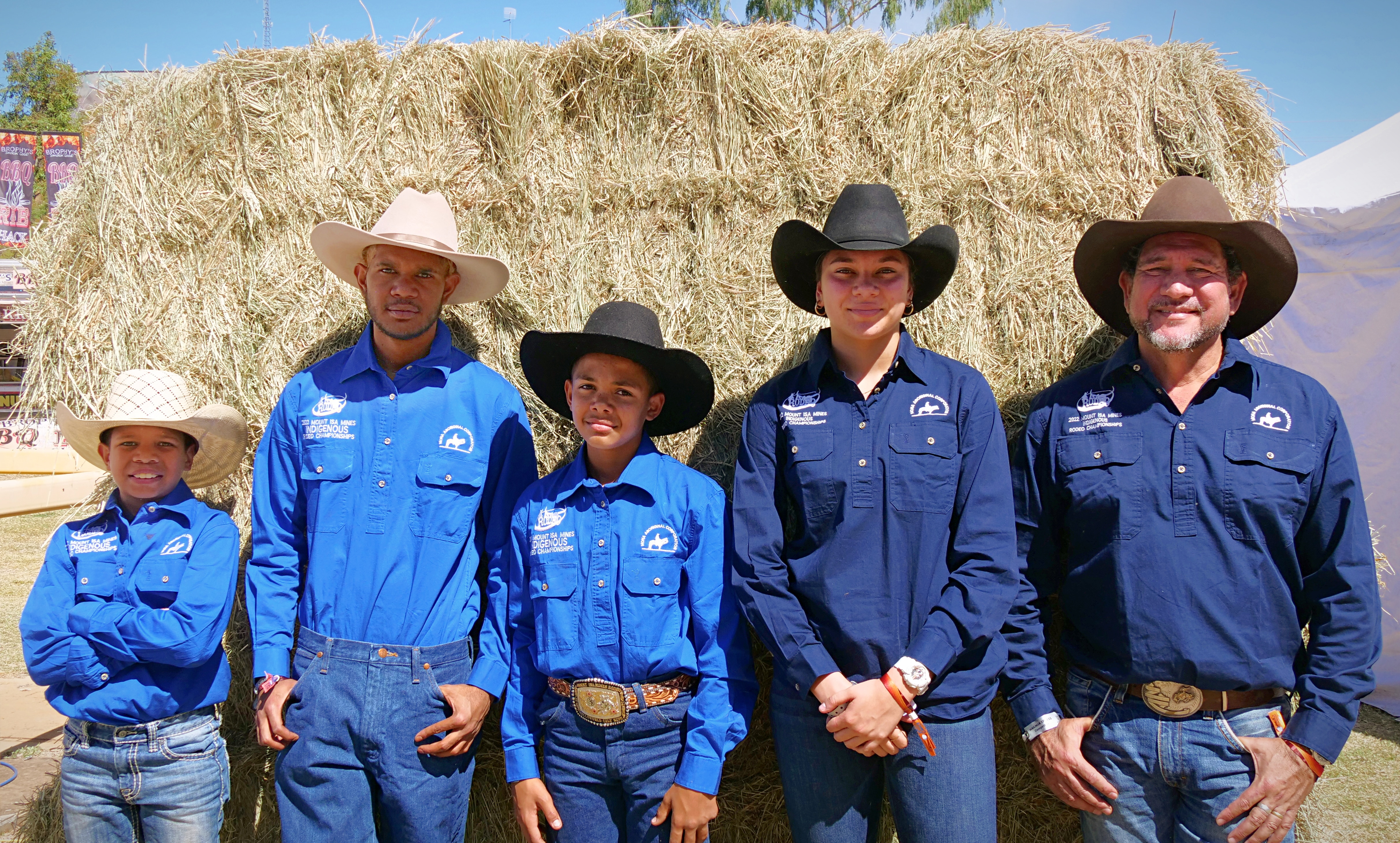 A group of young people and an older man, all wearing blue shirts and cowboy hats, stand in front of a hay bale