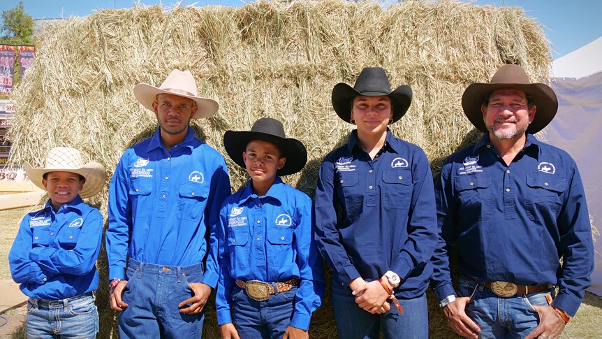 A group of young people and an older man, all wearing blue shirts and cowboy hats, stand in front of a hay bale