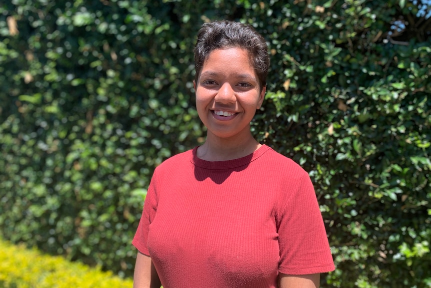 A young woman looks to the camera and smiles as she stands in front of a hedge, wearing a red t-shirt.