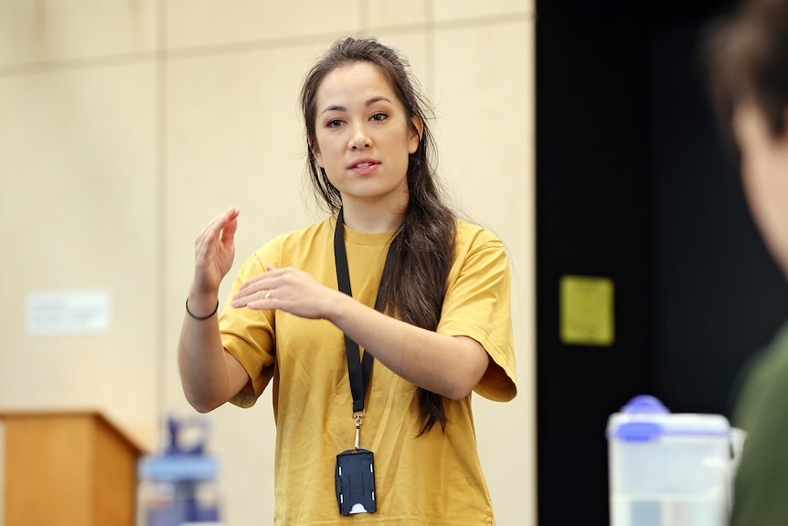 An Asian Australian woman in a yellow shirt stands, gesturing with her hands as she addresses people out of frame