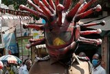 A man dressed in police uniform walks on an Indian street wearing a helmet with red spikes coming out of it.