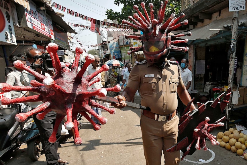 A man dressed in police uniform walks on an Indian street wearing a helmet with red spikes coming out of it.