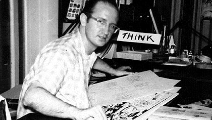 In a black and white photo, Steve Ditko sits at a desk with a pencil in his hand.