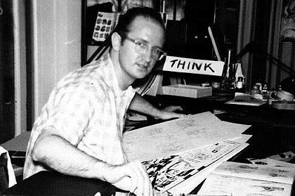 In a black and white photo, a man sits at a desk with a pencil in his hand.