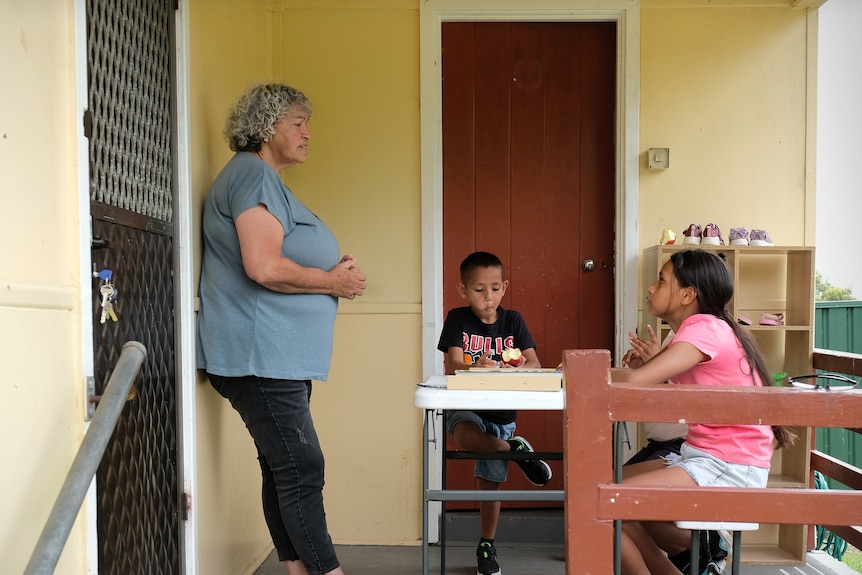 Two young kids are sitting at a table on a porch, studying and looking up at grandmother Jill.