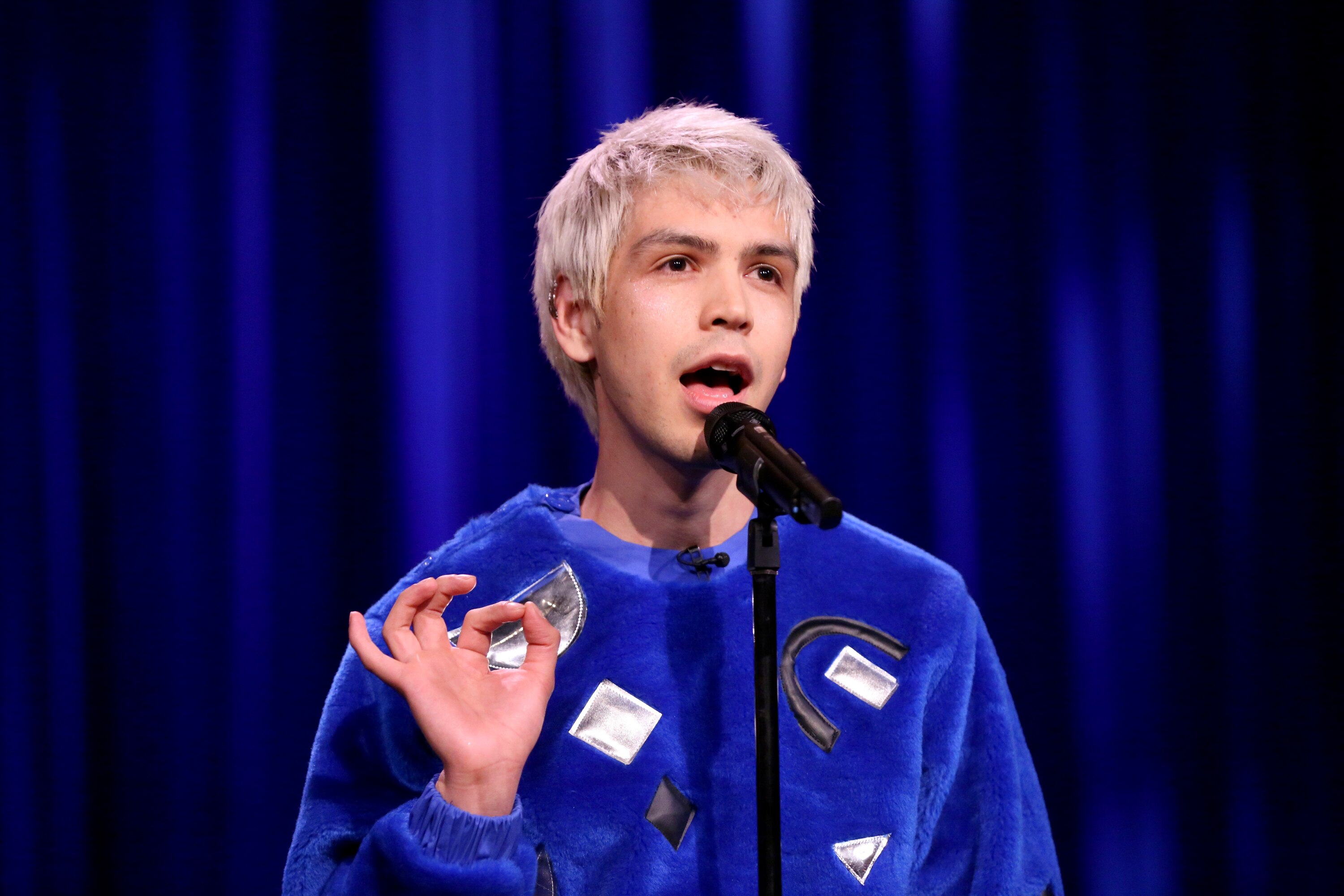 A man with bleached hair performs stand-up in front of a mic, wearing a blue velvet jumper with metallic shapes on it.