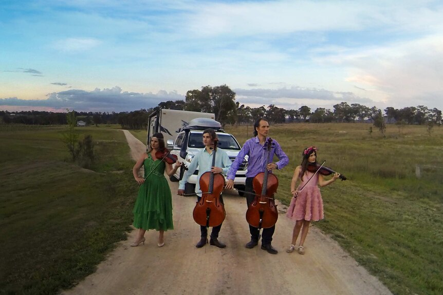 The Moir family stand with their instruments on an empty dirt road with their caravan behind them.