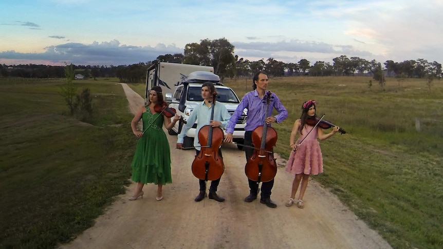 The Moir family stand with their instruments on an empty dirt road with their caravan behind them.