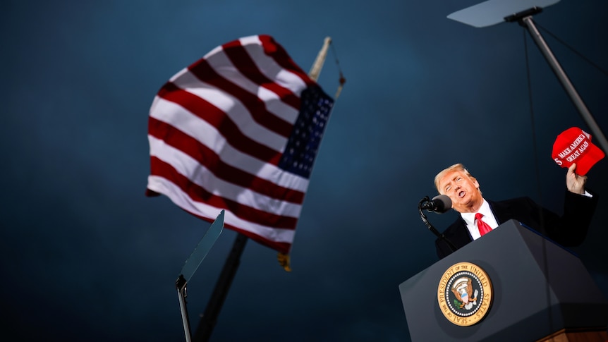 Donald Trump, seen from below, holds a red MAKE AMERICA GREAT AGAIN cap, beside a US flag against a stormy sky