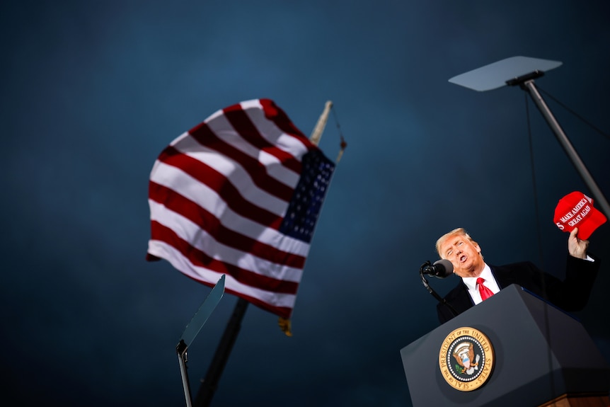 Donald Trump, seen from below, holds a red MAKE AMERICA GREAT AGAIN cap, beside a US flag against a stormy sky