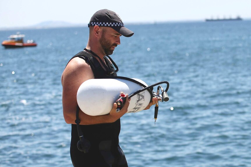 A side-on mid shot of a police diver carrying a white oxygen tank with the ocean in the background.