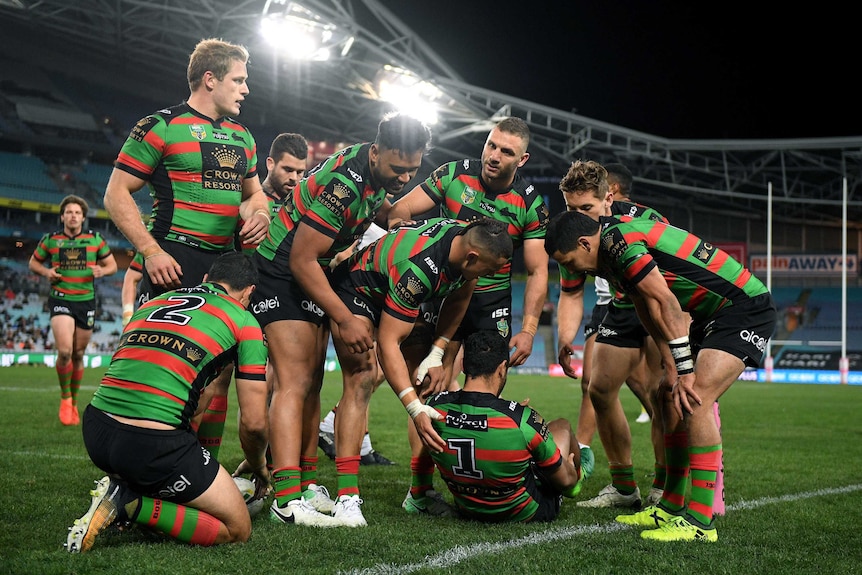 Alex Johnston sitting on the ground as Rabbitohs teammates surround him.