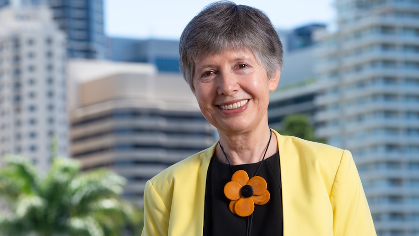 An image of Professor Lidia Morawska smiling wearing a yellow jacket, black blouse and orange flower necklace  buildings, tree