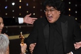 Bong Joon Ho opens his mouth in joy as he looks at Jane Fonda, seen from behind, holding his Oscar out to him.