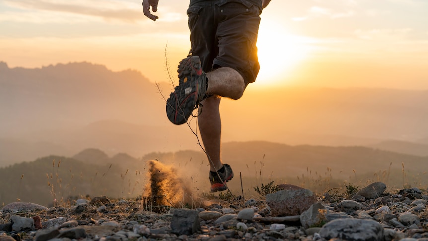 lower half of man in running pose, trail shoes kicking up dust amongst pebbles, mountains and sunrise in background